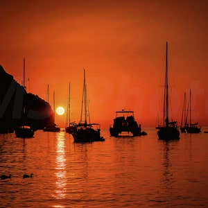 Puede incluir: Silueta de veleros y otras embarcaciones sobre agua tranquila al atardecer. El sol es un orbe naranja brillante, que se refleja en el agua. Una pequeña isla y un faro son visibles en el fondo.
