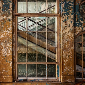 Puede incluir: Vista interior de un edificio en ruinas con pintura descascarillada en tonos amarillos, azules y marrones. Una gran ventana revela una escalera con barandillas ornamentadas. También se ven ventanas arqueadas y circulares.