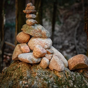 Puede incluir: Un cairn de piedra equilibrado, compuesto por rocas naturales de varios tamaños y formas, se alza sobre una base musgosa. Las piedras varían en color desde el marrón claro hasta el marrón rojizo, con un fondo borroso de bosque.