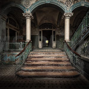 Puede incluir: Interior de un edificio antiguo y en ruinas con una gran escalera. Las paredes muestran pintura descascarillada en tonos azules y grises. Dos grandes columnas flanquean la escalera, creando una atmósfera de abandono.