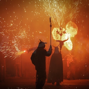 Puede incluir: Escena nocturna con figuras silueteadas en ropa oscura, algunas con cuernos, sosteniendo antorchas y fuegos artificiales. El fondo está lleno de explosiones y chispas de fuego, iluminado por luz naranja y amarilla, creando una atmósfera dramática.