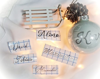 May include: A clear glass Christmas ornament with the name "Kiar" in silver script. Several name decals, including "Alina", "David", "Tobias", "Maximilia", and "Alexander", are displayed. A small white wooden sled and a pine cone are also visible.
