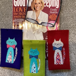 May include: Three felt eyeglass cases with embroidered cat designs. The cases are blue, green and red. The blue case has a cat with a teal bow tie and a floral patterned tail. The green case has a cat with a teal bow tie and a floral patterned tail. The red case has a cat with a white bow tie and a red and white star patterned tail.