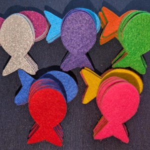 May include: A set of ten felt fish shapes in various colours, including silver, purple, blue, orange, green, red, and pink. Each fish is made of multiple layers of felt, creating a dimensional effect.