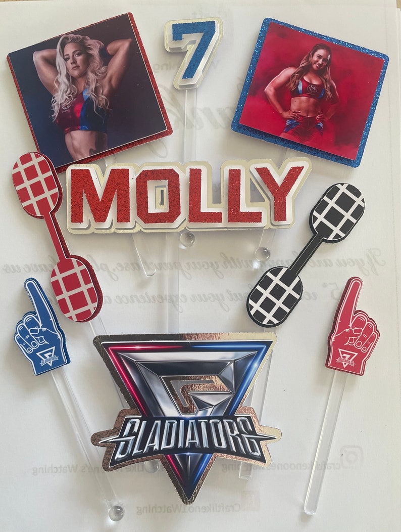 Puede incluir: Un conjunto de adornos para pasteles con una luchadora, el n&uacute;mero 7, la palabra "MOLLY", un logotipo de gladiador y dedos de espuma. La luchadora lleva un atuendo rojo, blanco y azul. El n&uacute;mero 7 es azul. La palabra "MOLLY" es roja. El logotipo del gladiador es plateado, azul y rojo. Los dedos de espuma son rojos y azules.