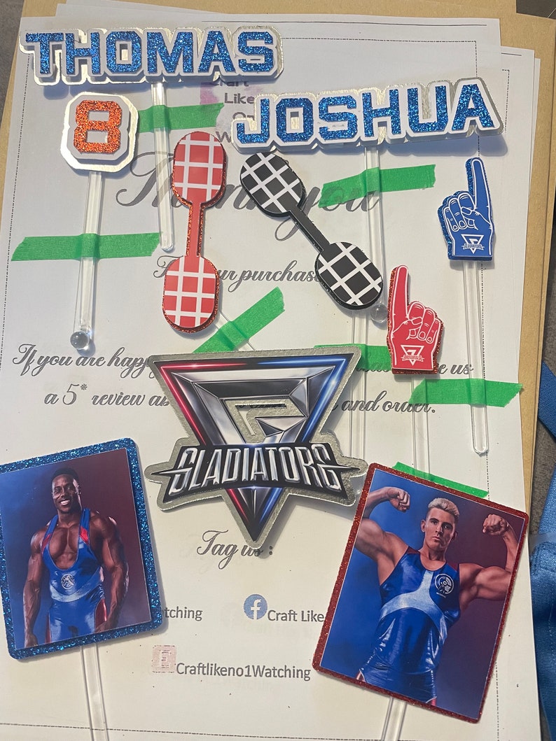 Puede incluir: Un conjunto de adornos para pasteles con los nombres "Thomas" y "Joshua" con el n&uacute;mero "8" en medio. Los adornos est&aacute;n decorados con purpurina y presentan un esquema de colores rojo, blanco y azul. Los adornos tambi&eacute;n est&aacute;n decorados con im&aacute;genes de luchadores y la palabra "Gladiators".