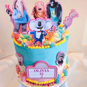 May include: A birthday cake with blue icing and colourful buttercream decorations. The cake is decorated with character cutouts from the film "Sing", including a koala, elephant, and pig. The cake has a sign that says "Olivia is 2".