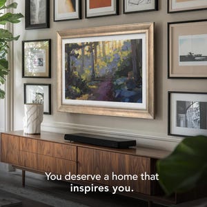 Puede incluir: Una escena de sala de estar con una gran obra de arte enmarcada que representa un bosque, rodeada de imágenes enmarcadas más pequeñas. Un mueble de madera oscura con una barra de sonido negra se encuentra debajo de la obra de arte. El texto "You deserve a home that inspires you." está en la parte inferior.