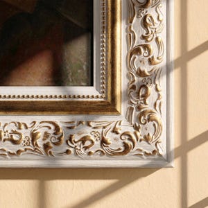 Könnte beinhalten: Nahaufnahme eines dekorativen Bilderrahmens. Der Rahmen hat eine weiße Basis mit goldenen Akzenten und verzierten, geschnitzten floralen Designs. Der Innenrand hat eine goldene Perlenverzierung. Der Rahmen wirft teilweise einen Schatten auf eine beige Wand.