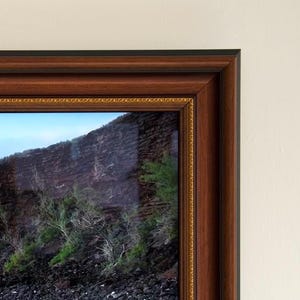 Pode incluir: Fotografia de paisagem emoldurada, com uma moldura de madeira castanha escura e uma borda interna dourada. A foto mostra uma costa rochosa, uma montanha e um céu azul.