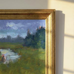 Pode incluir: Pintura de paisagem emoldurada com uma moldura dourada ornamentada. A obra retrata um campo verde, um corpo de água e uma floresta sob um céu azul nublado. A pintura é iluminada por luz natural.