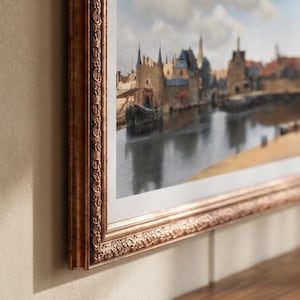 Op de afbeelding: Een ingelijste prent van een Europese stadsgezicht met een kanaal en gebouwen. De lijst is versierd, met een bronzen afwerking. Het kunstwerk toont een gedetailleerde scène met boten, water en architectuur, die geschiedenis en cultuur oproept.