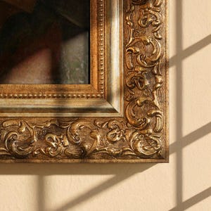 Pode incluir: Close-up de uma moldura dourada com entalhes detalhados. A moldura, de forma retangular, apresenta uma borda decorativa e uma superfície texturizada. Está colocada contra uma parede neutra com sombras.