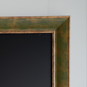 Op de afbeelding: Een rechthoekig schoolbord met een verweerde groen en goud geschilderde houten lijst. De lijst heeft een getextureerd uiterlijk, met de groene verf die op sommige plaatsen door het goud heen schijnt. Het schoolbordopper-vlak is zwart.