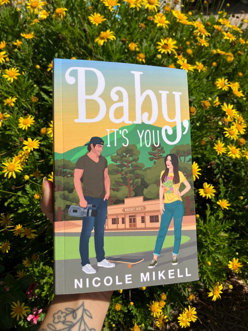 May include: A book cover with the title "Baby, It's You" and the author's name, Nicole Mikell. The cover art depicts a man and woman standing in front of a building. The background features a landscape with trees and a sunset.