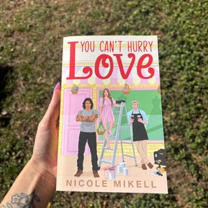 Pu&ograve; includere: Un libro tascabile intitolato "YOU CAN'T HURRY LOVE" di Nicole Mikell. La copertina presenta illustrazioni di tre persone, un edificio rosa e una scala. Il titolo &egrave; in grandi lettere rosse.