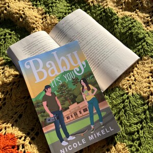 May include: A paperback book titled "Baby It's You" by Nicole Mikell, resting on top of an open book. The cover features illustrations of a man and woman. The books are placed on a colorful, textured blanket with orange, green, and yellow hues.
