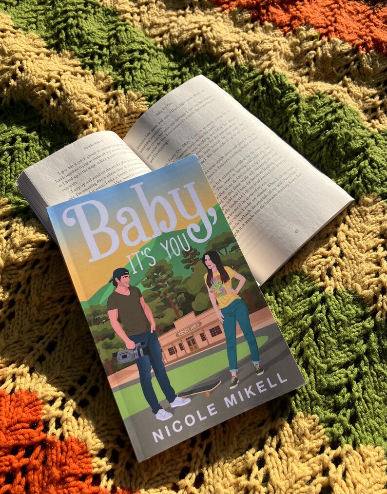 May include: A paperback book titled "Baby It's You" by Nicole Mikell, resting on top of an open book. The cover features illustrations of a man and woman. The books are placed on a colorful, textured blanket with orange, green, and yellow hues.