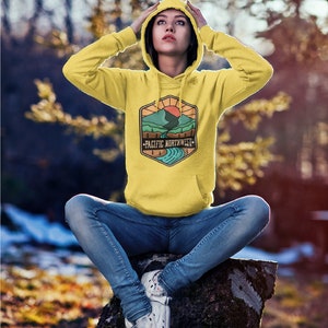 May include: A yellow hoodie with a graphic of a mountain range and a river with the text "Pacific Northwest" on it. The person wearing the hoodie is sitting on a tree stump in a forest.