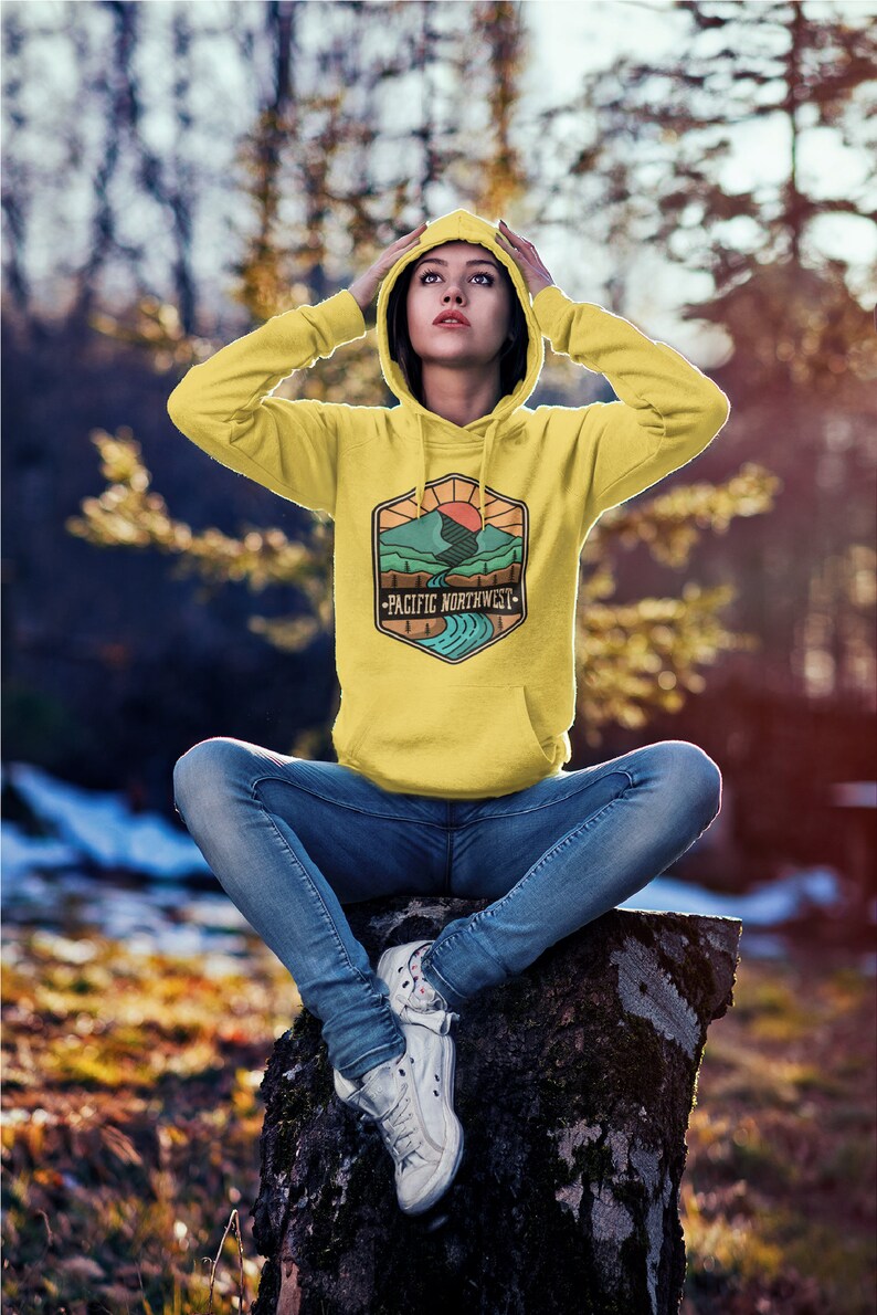 May include: A yellow hoodie with a graphic of a mountain range and a river with the text "Pacific Northwest" on it. The person wearing the hoodie is sitting on a tree stump in a forest.