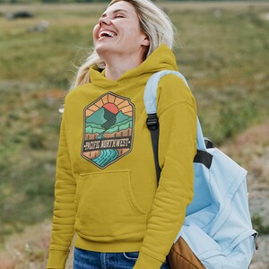 May include: A mustard yellow hoodie with a graphic of a mountain range, a river, and a sun. The graphic is in a hexagon shape with the text "Pacific Northwest" below it.