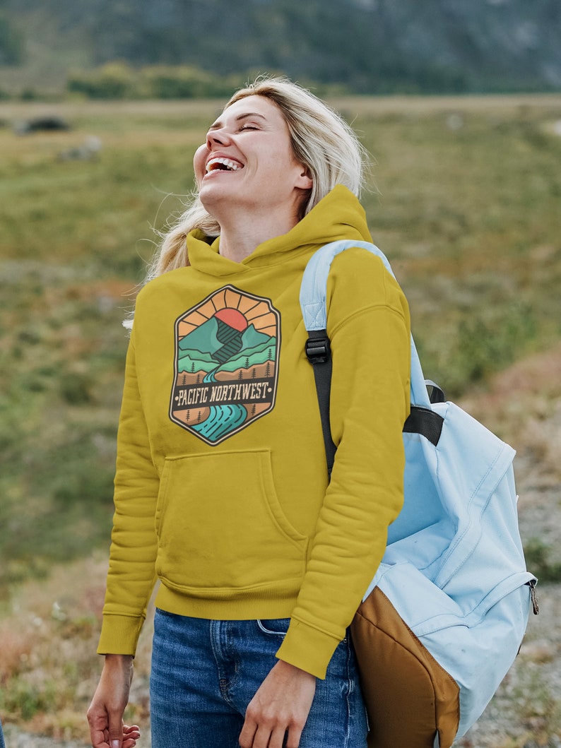 May include: A mustard yellow hoodie with a graphic of a mountain range, a river, and a sun. The graphic is in a hexagon shape with the text "Pacific Northwest" below it.