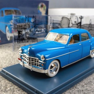 May include: A bright blue vintage car model, featuring a classic design with chrome detailing and white-walled tyres. The model is presented on a matching blue base, with a scale of 1:24. The car's features are meticulously crafted.