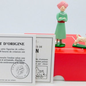 Könnte beinhalten: Ein Herkunftszertifikat für eine Sammlerfigur. Das Zertifikat ist auf weißem Papier mit schwarzem Text gedruckt. Der Text lautet "Certificat d'Origine". Das Zertifikat befindet sich neben einer roten Schachtel mit einer grünen Figur einer Frau, die einen rosa Turban trägt, und einem weißen Hund, der einen rosa Turban trägt. Die Figur steht auf einem grünen Sockel. Der Text auf der Schachtel lautet "Les Aventures".