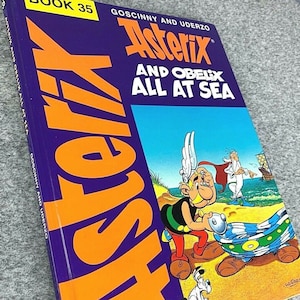 Könnte beinhalten: Ein lila und orangefarbenes Buch mit dem Titel "Asterix und Obelix auf hoher See" mit einer Comic-Illustration von Asterix und Obelix auf dem Cover. Das Buch ist Teil einer Reihe und trägt die Nummer "Buch 35".