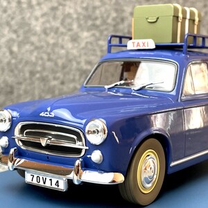 May include: A blue vintage taxi model with a luggage rack on the roof, holding two green suitcases. The car has a "TAXI" sign and a license plate that reads "70V14". The model car is a Peugeot 403.