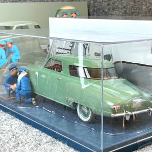 Könnte beinhalten: Ein hellgrünes Oldtimer-Automodell in einer transparenten Vitrine. Das Modell hat Weißwandreifen, einen roten Innenraum und Chromakzente. Miniaturfiguren von Mechanikern in blauen Uniformen sind um das Auto herum positioniert. Die Vitrine hat einen blauen Sockel und den Text "JEUDI 18 AOUT".