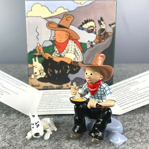 Könnte beinhalten: Ein Figurenset mit einer Comicfigur eines Cowboys, der auf einem Felsen sitzt, aus einer Pfanne mit einem Löffel isst und einen weißen Hund, der sich hinlegt. Die Figur trägt ein blau-weiß kariertes Hemd, ein rotes Halstuch und einen braunen Cowboyhut. Das Set wird mit einer Schachtel geliefert, die ein Comicbild des Cowboy-Charakters zeigt.