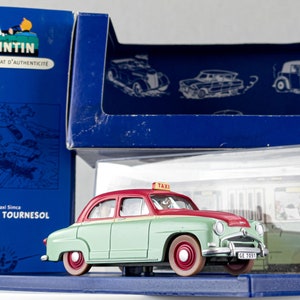 May include: A model of a vintage taxi car, red and green, with the text "TAXI" on top. The car is in a clear display case, with a blue box featuring Tintin artwork and the text "CERTIFICAT D'AUTHENTICITE".