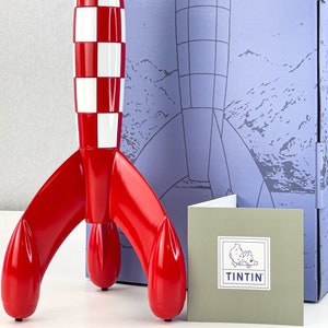 May include: A red and white rocket ship model with a checkered pattern on the body. The rocket is on a blue and white box with a smaller box that says "Tintin" on it.