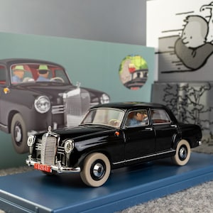 Puede incluir: Un modelo de coche Mercedes-Benz negro con neum&aacute;ticos blancos, expuesto sobre una base azul. El modelo presenta una parrilla cromada detallada y una matr&iacute;cula roja. La caja muestra un coche similar y una ilustraci&oacute;n de dibujos animados.