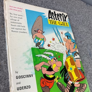 May include: A book cover featuring a cartoon illustration of Asterix, a Gaul warrior, fighting a Roman soldier. The book title is "Asterix the Gaul" and the authors are Goscinny and Uderzo.