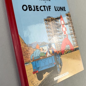 May include: A hardcover book titled "OBJECTIF LUNE" with a red spine. The cover features a blue jeep with characters, a red and white rocket, and the text "HERGE - LES AVENTURES DE TINTIN". The book is published by Casterman.