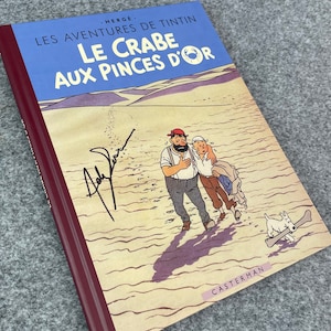 May include: A vintage hardcover book titled "Les Aventures de Tintin Le Crabe aux Pinces d'Or" by Herge. The cover features a cartoon illustration of Tintin and Captain Haddock walking on a sandy beach with Snowy the dog.