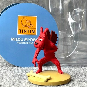 Puede incluir: Una figura de resina roja de Milú, el perro de Tintín, vestido de diablo con alas y sosteniendo un hueso. La figura está sobre una base de color beige y viene en un contenedor de plástico transparente. El embalaje incluye el logotipo de Tintín y el texto "Milou Mi-Dém  figurine résine".