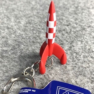 Könnte beinhalten: Ein roter und weißer Raketen-Schlüsselanhänger mit einem silbernen Ring und einem blauen Anhänger mit dem Wort "Tintin" und einem Comicbild von Tintin.