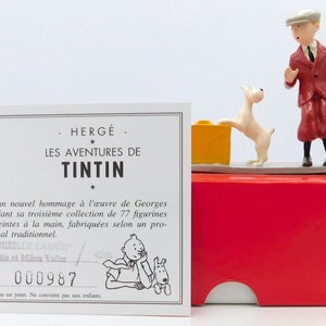 Könnte beinhalten: Eine weiße Karte mit schwarzem Text lautet "HERGÉ LES AVENTURES DE TINTIN" und "PIXI rend un nouvel hommage à l'œuvre de Georges Rémi en créant sa troisième collection de 77 figurines en métal, peintes à la main, fabriquées selon un procédé artisanal traditionnel." Die Karte trägt auch die Nummer "000987". Eine rote Box mit einem weißen Ausschnitt auf der Vorderseite enthält eine kleine weiße Hundefigur und eine kleine Figur eines Mannes in rotem Anzug und grauem Hut. Der Mann schaut auf den Hund, der auf einem gelben Block steht.