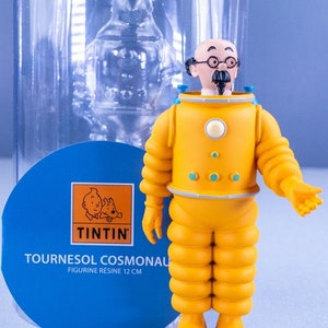 May include: A yellow and white figurine of Professor Calculus from the Tintin comics wearing a yellow space suit. The figurine is 12 cm tall and is made of resin. The figurine is on a tan base and is in a clear plastic box. The box has a blue label with the Tintin logo and the text "Tournesol Cosmonaut Figurine R&eacute;sine 12 cm".