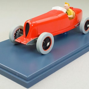 Pu&ograve; includere: Modello di auto da corsa rossa vintage con figura di pilota, esposto su una base rettangolare blu. L'auto ha ruote grigie con raggi neri e un roll bar bianco. Il pilota indossa una camicia gialla.