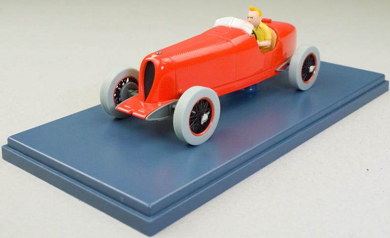 Pu&ograve; includere: Modello di auto da corsa rossa vintage con figura di pilota, esposto su una base rettangolare blu. L'auto ha ruote grigie con raggi neri e un roll bar bianco. Il pilota indossa una camicia gialla.