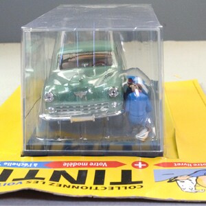 Pu&ograve; includere: Un modello di auto d'epoca verde in una vetrina di plastica trasparente. Il modello &egrave; una replica dettagliata, con paraurti anteriore e fari argentati. Una piccola figura in uniforme blu &egrave; accanto all'auto. Il modello &egrave; su una carta gialla.