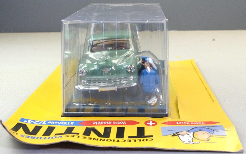 Pu&ograve; includere: Un modello di auto d'epoca verde in una vetrina di plastica trasparente. Il modello &egrave; una replica dettagliata, con paraurti anteriore e fari argentati. Una piccola figura in uniforme blu &egrave; accanto all'auto. Il modello &egrave; su una carta gialla.