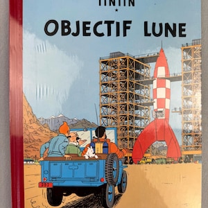 May include: A hardcover comic book titled "OBJECTIF LUNE" by Herg&eacute;. The cover features a blue jeep with Tintin, Captain Haddock, and Snowy, with a red and white rocket in the background. The book is sealed in plastic.