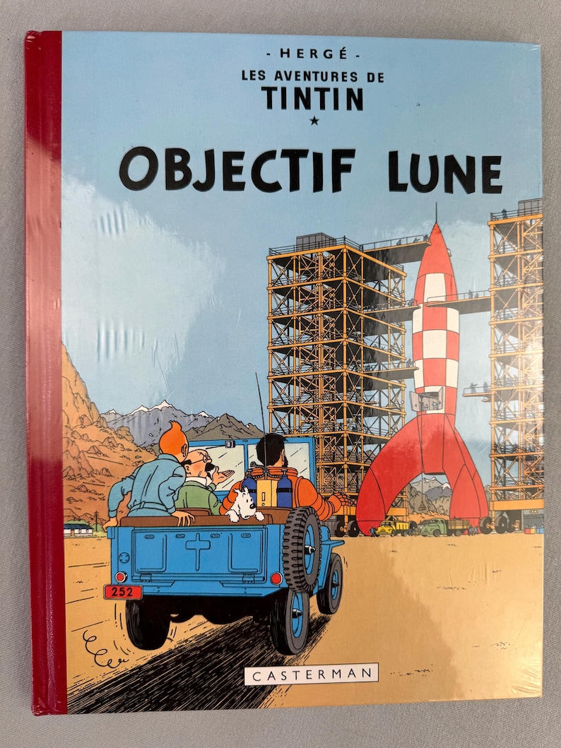 May include: A hardcover comic book titled "OBJECTIF LUNE" by Herg&eacute;. The cover features a blue jeep with Tintin, Captain Haddock, and Snowy, with a red and white rocket in the background. The book is sealed in plastic.