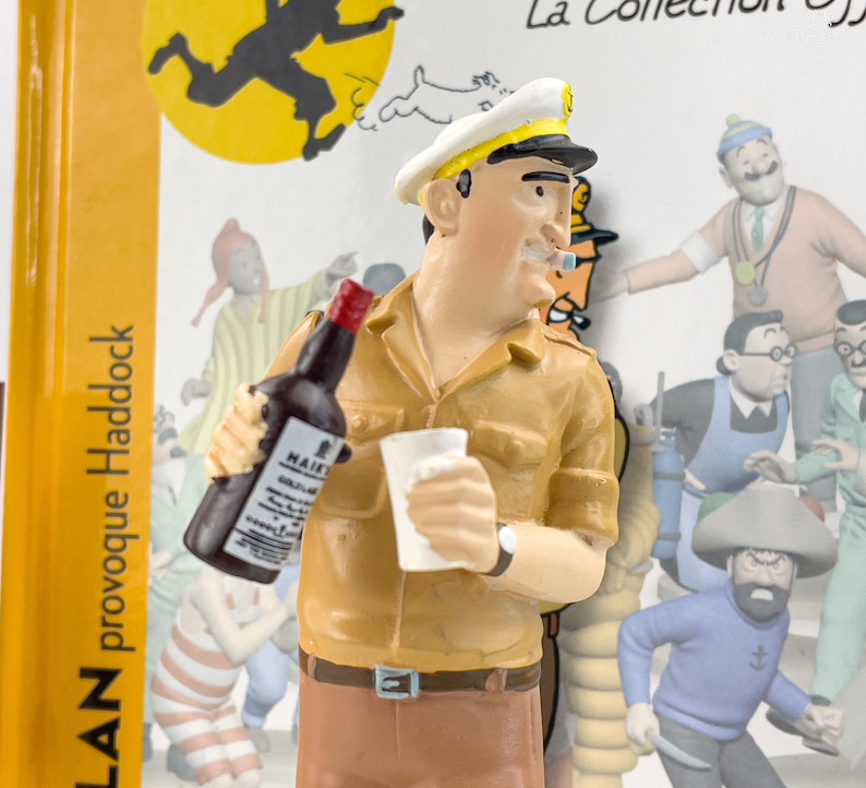 May include: A figurine of a cartoon character wearing a captain's hat and holding a bottle of alcohol and a cup. The character is wearing a brown shirt and pants. The figurine is part of a collection of cartoon characters.