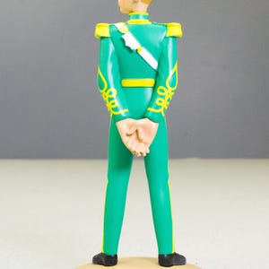 May include: A figurine of a man in a green uniform with yellow accents and a white sash. The figure has blond hair and is standing on a tan circular base. The uniform features decorative yellow scrollwork.